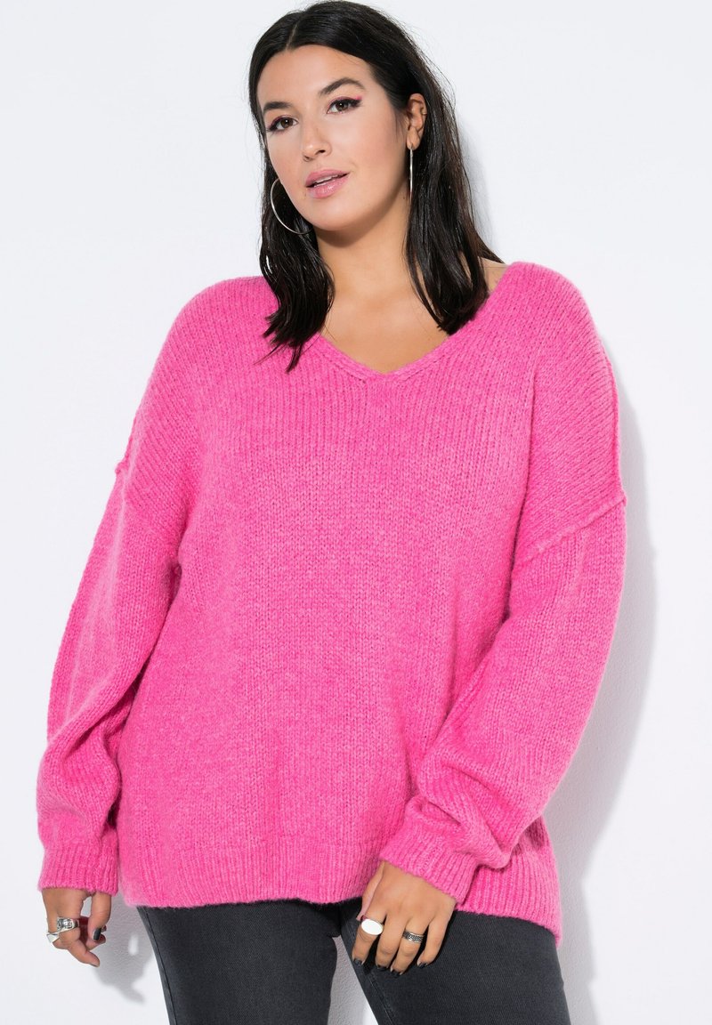 Oversized różowy sweter o ribbowanym wzorze, z dekoltem w kształcie litery V i długimi rękawami. W zestawieniu z ciemnoszarymi dżinsami.