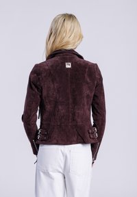 Burgundská semišová bunda s límcem, s kovovými akcenty ve stříbrné barvě a nastavitelnými bočními detaily, spárovaná s bílými denimovými kalhotami.