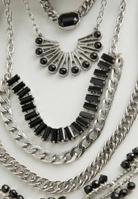 Slojevite ogrlice s srebrnim lancima i crnim akcentima, uključujući pravokutne i okrugle perle, koje ističu raznolike dizajne i teksture.