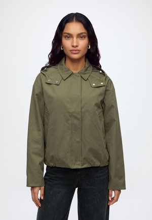 Femme aux longs cheveux foncés portant une veste à capuche verte et un pantalon noir, debout devant un fond clair uni.