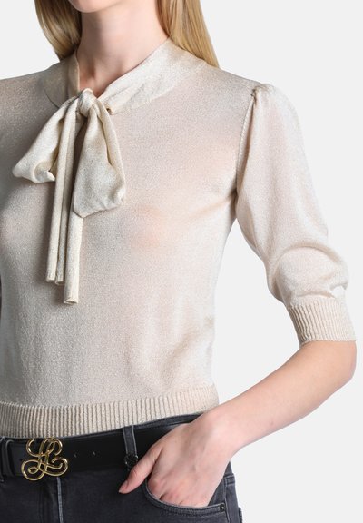 Femme portant un haut en maille beige brillant avec des manches bouffantes et un nœud noué au cou, associé à un pantalon noir et une ceinture noire avec une boucle dorée ornée.