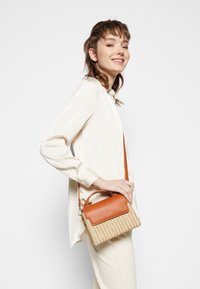 Jeune femme en blouse crème et pantalon portant un petit sac tissé avec rabat en cuir brun et bandoulière, souriant de profil.