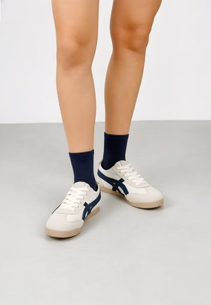 Piernas con zapatillas blancas con rayas azul marino y suelas beige combinadas con calcetines navy de caña media, de pie sobre un suelo gris claro.