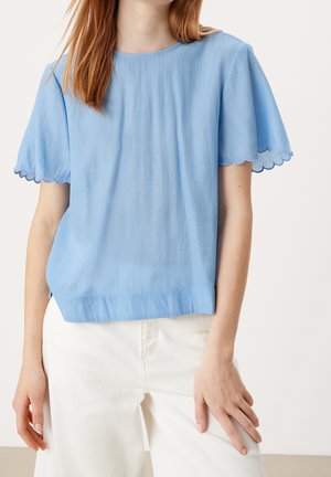 Femme portant un haut texturé bleu clair à manches courtes avec des bords festonnés et un pantalon blanc taille haute sur un fond uni.