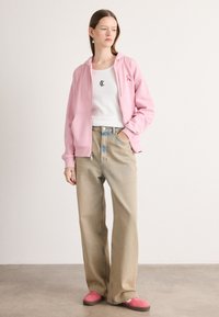Felpa rosa con tasche frontali, canotta bianca con logo nero, jeans a gamba larga azzurri e sneakers rosa con suola scura.