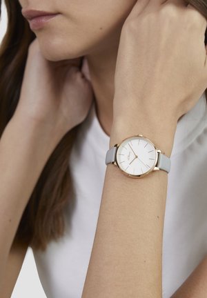 Runde Uhr mit weißem Zifferblatt, goldfarbenem Gehäuse und schlankem grauem Silikonarmband. Schlichte Stundenmarkierungen und minimalistisches Design.