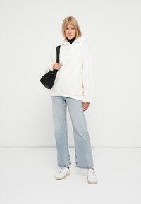 Veľká smotanová mikina s predným logom, kombinovaná s svetlomodrými rozšírenými džínsami a bielymi teniskami. Doplnkom je čierna crossbody taška.