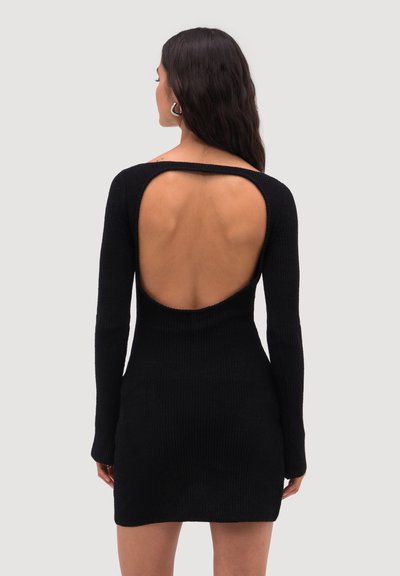 Femme aux longs cheveux noirs portant une robe noire à manches longues en maille avec une grande découpe ovale dévoilant le haut de son dos.