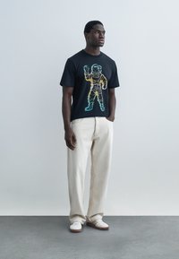 Uomo in piedi con le mani nelle tasche che indossa una maglietta nera con una grafica colorata di un astronauta, pantaloni chiari e sneaker bianche.