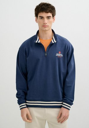 Joven hombre con sudadera azul marino con cremallera hasta el pecho, puños y dobladillo con rayas blancas y el logo de "Whitehall Rowing Club" en el pecho.
