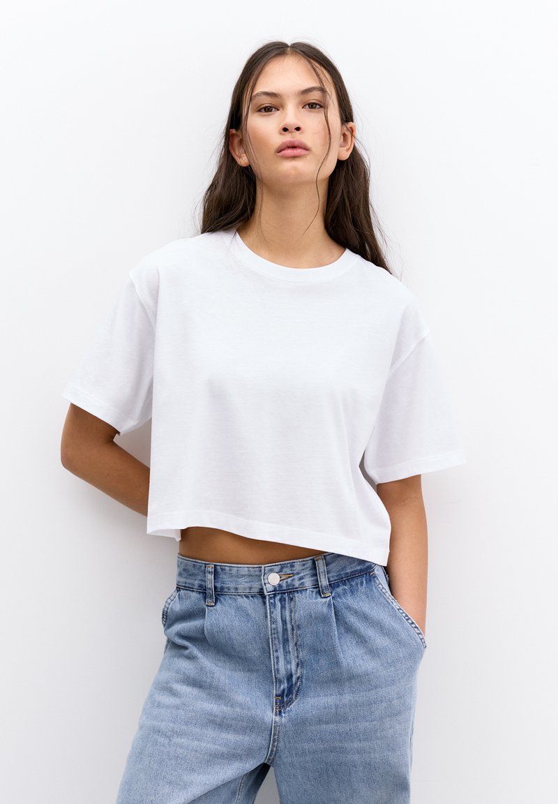 Tricou alb scurt, cu mâneci scurte, din bumbac moale. Asortat cu blugi de talie înaltă, de culoare albastru deschis, cu închidere prin buton și buzunare laterale.