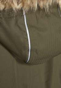 La tela de la chaqueta color verde oliva presenta un patrón de espiga, con un ribete plateado a lo largo de la capucha y un borde de piel en el cuello.