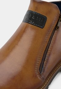 Brązowy skórzany botek za kostkę o gładkiej fakturze, z bocznym zamkiem błyskawicznym i czarną metką marki. Posiada zaokrąglony czubek i kontrastowe szwy.