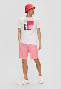 Witte t-shirt met een kleurblokontwerp met roze en blauwe grafische elementen, gecombineerd met roze shorts en een rode bucket hat met een patroon.