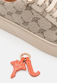 Beige sneaker featuring a textured upper with a subtle pattern and a cream rubber sole. Accompanied by an orange keychain with a floral design.