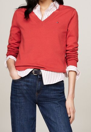 Vrouw draagt een koraalkleurige V-hals trui over een wit gestreept overhemd met opgerolde mouwen, gecombineerd met donkerblauwe jeans en een zwarte riem.