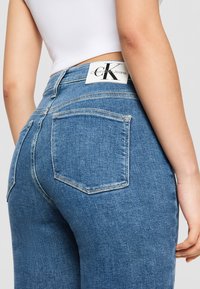 Blue denim jeans with a high waist, featuring two back pockets and a white label with “Calvin Klein Jeans” logo at the waistband.