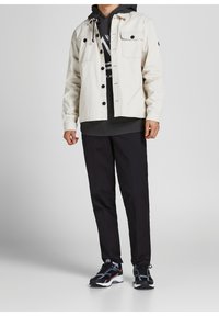 Chaqueta beige clara de botones con dos bolsillos frontales sobre una sudadera con capucha gris oscuro, combinada con pantalones negros y zapatillas deportivas negras.