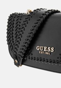 Borsa a tracolla in pelle nera con dettagli intrecciati, hardware dorato e logo impresso "GUESS EST 1981". Profilo piatto con design strutturato.