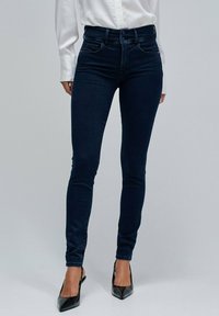 Kvinna klädd i mörkblå skinny jeans med dubbelknäppning, vit långärmad skjorta och svarta pumps med spetsig tå på en enfärgad bakgrund.