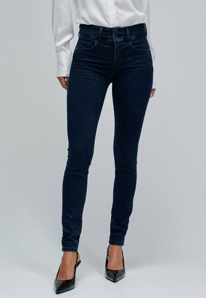 Mujer vistiendo vaqueros skinny azul oscuro con cintura de doble botón, camiseta blanca de manga larga y tacones negros de punta en un fondo liso.