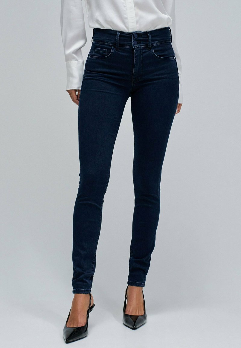 Kvinna klädd i mörkblå skinny jeans med dubbelknäppning, vit långärmad skjorta och svarta pumps med spetsig tå på en enfärgad bakgrund.