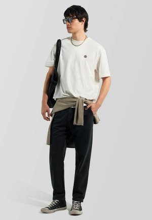 Junger Mann trägt weißes Grafik-T-Shirt, schwarze Hose, schwarze Sneakers, Sonnenbrille, mit beigefarbenem Pullover um die Taille gebunden, trägt schwarze Tasche.