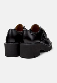 Chaussures en cuir noir avec une semelle noire épaisse, une forme arrondie et un détail de boucle visible. La doublure intérieure est marron.