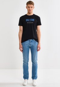 Mladý muž oblečený v černém tričku s nápisem "BIG STAR", světle modrých úzkých džínách a bílých teniskách, stojící před jednobarevným bílým pozadím.