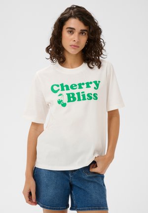 Woman with curly hair wears white T-shirt with green "Cherry Bliss" text and cherry graphic, paired with blue denim shorts.