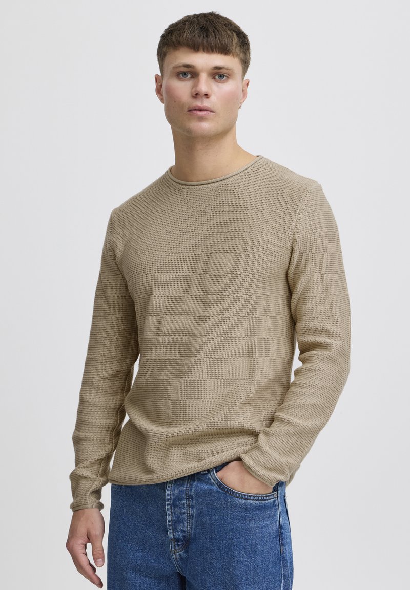 Ung mand med kort brunt hår iført en beige tekstureret sweater og blå jeans, stående med den ene hånd i lommen mod en ensfarvet baggrund.