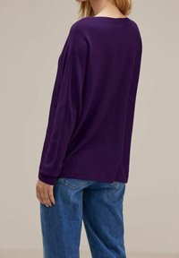 Kvinna iförd en lös lila långärmad topp och blå jeans, visad bakifrån mot en enkel bakgrund.