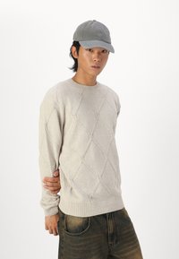 Beige knitted sweater with diamond pattern and ribbed neckline. Worn with dark jeans. Grey baseball cap completes the look.