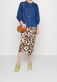 Donna che indossa una blusa blu con bottoni, una gonna midi con stampa leopardata, tacchi a blocco verdi e porta una borsa a tracolla marrone.