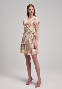 Leichtes, pastellfarbenes Paisley-Wickelkleid mit Rüschensaum, kurzen Ärmeln und einem Bindegürtel. Trägt rosa High Heels. Glatte Stofftextur.