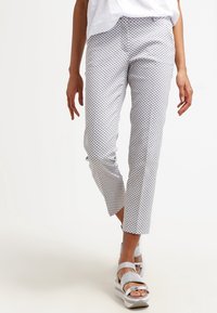 Kobieta idąca w białych spodniach z szarym wzorem w diamenty i białych sandałach na platformie, z częściowo widoczną białą górą.