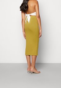 Olivově zelená žebrovaná midi sukně s přiléhavým střihem, vybavená rozparkem vzadu. V kombinaci s průhlednými sandály na podpatku. Zvýrazněný zadní pohled.