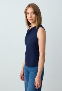 Top senza maniche navy con scollo rotondo e una leggera lucentezza, abbinato a jeans blu medio. Il top ha una texture liscia e un design minimalista.