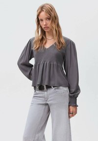Jeune femme aux cheveux blonds ondulés portant un haut smocké gris à manches longues et un jean gris clair, posant avec une main sur la hanche.