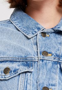 Näruppbild av en ljusblå denimjacka med krage och bröst, med metallknappar märkta "GUESS" och synliga söm detaljer.