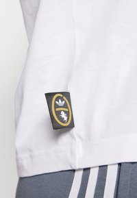 Camiseta blanca de algodón con una etiqueta negra y dorada que presenta el logo de Adidas y un gráfico de dinosaurio, situado cerca del dobladillo.