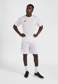 T-shirt et shorts de sport blancs avec des rayures noires. Le t-shirt présente un logo et a des manches courtes. Tissu texturé, coupe sportive.