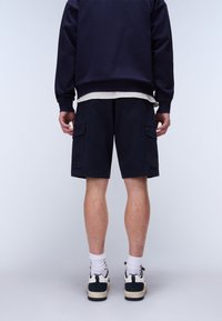 Person trägt marineblaue Cargo-Shorts, marineblauen Pullover, weiße Socken und schwarz-weiße Turnschuhe und steht vor einem hellen Hintergrund.