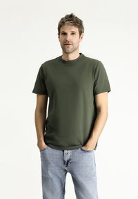T-shirt verde oliva a maniche corte con scollatura rotonda, realizzato in tessuto morbido. Indossato con jeans in denim azzurri chiari. Design semplice, senza motivi.