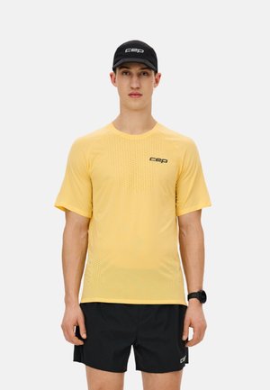 Man in yellow short-sleeve sports shirt and black shorts, wearing black cap and wristwatch, standing against light gray background.