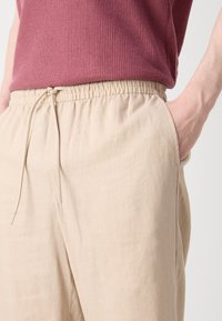 Beige linbukser med elastisk talje og snøre detaljer; sidelommer synlige. Båret over en teksturert mørkerød topp.