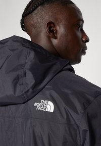 Svart nylonjacka med huva, med The North Face-logotyp i vitt. Slät textur och enkelt design, visar axeln och ryggen.