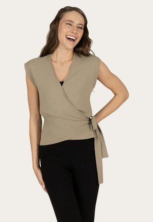 Frau lächelt, trägt ein ärmelloses beiges Wickeltop, das in der Taille gebunden ist, und schwarze Hosen, steht mit einer Hand auf der Hüfte.