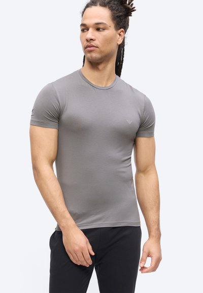 Άνδρας με μακριά dreadlocks που φοράει εφαρμοστό γκρι κοντομάνικο μπλουζάκι και μαύρο παντελόνι, κοιτάζοντας προς τα αριστερά σε απλό φόντο.