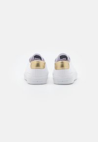 Sneakers bianche con una finitura liscia, caratterizzate da un accento metallico oro sul tallone e una suola in gomma testurizzata. Design minimale.